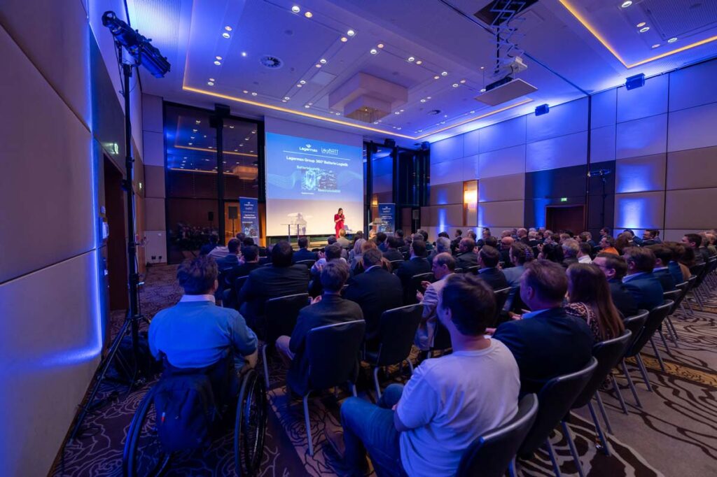 Eine Rednerin spricht vor einem aufmerksam zuhörenden Publikum in blau beleuchtetem Konferenzraum
