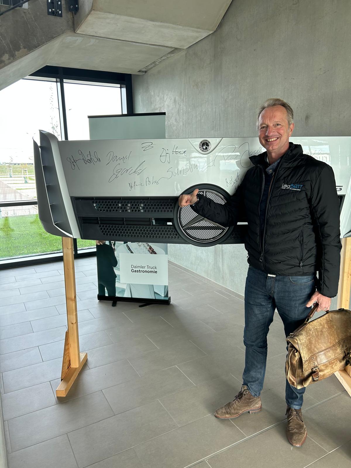 Ein lächelnder Mann in schwarzer Jacke steht neben einer signierten Lkw-Front in modernem Raum