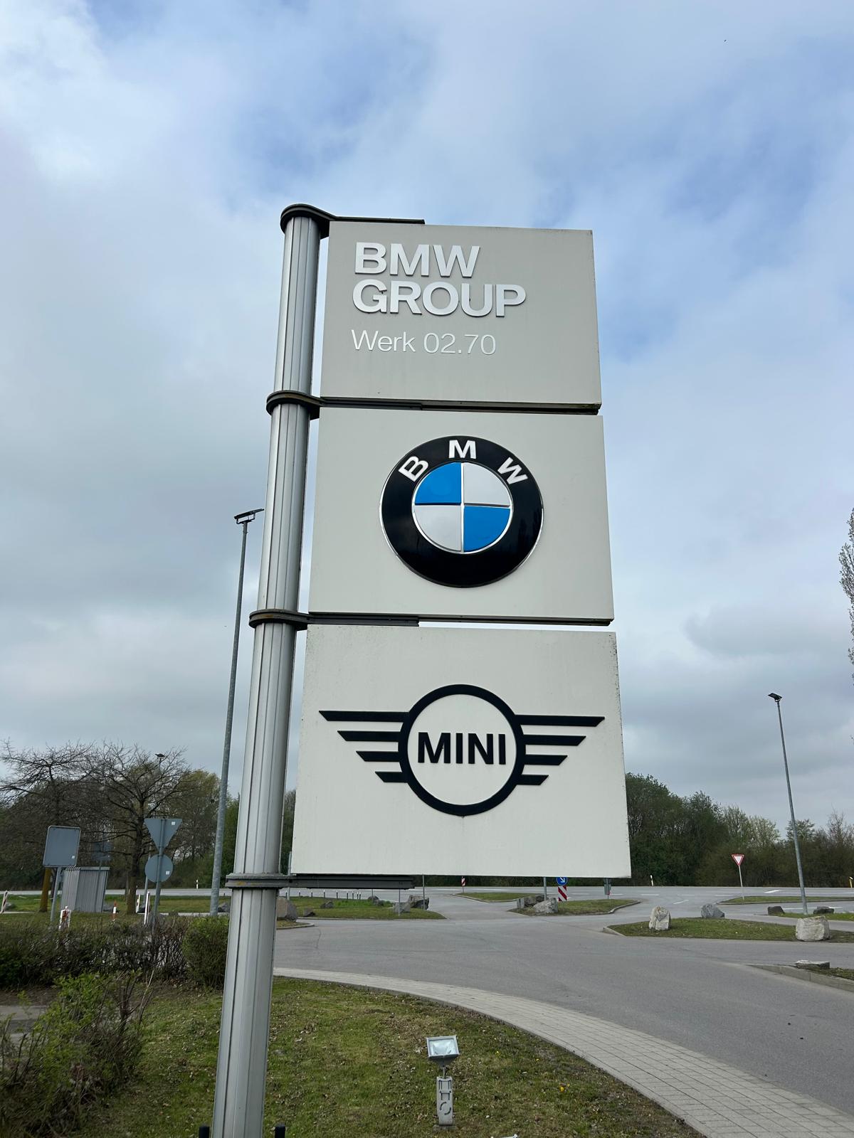 Ein Schild mit BMW und MINI Logos steht an einer Straße vor grünem Hintergrund