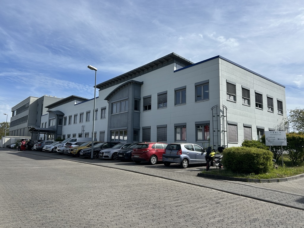 Graues Bürogebäude mit vielen Autos davor unter blauem Himmel und grüner Bepflanzung