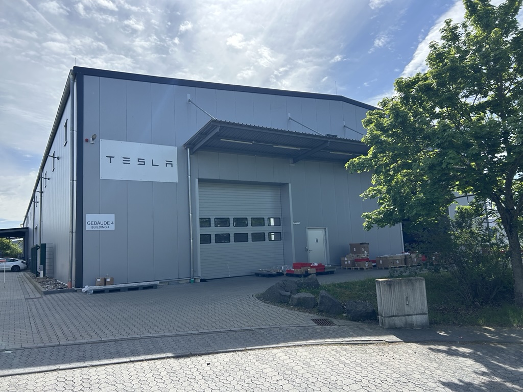 Graues Tesla Gebäude mit großer Garage und mehreren Paketen, Baum vorne rechts