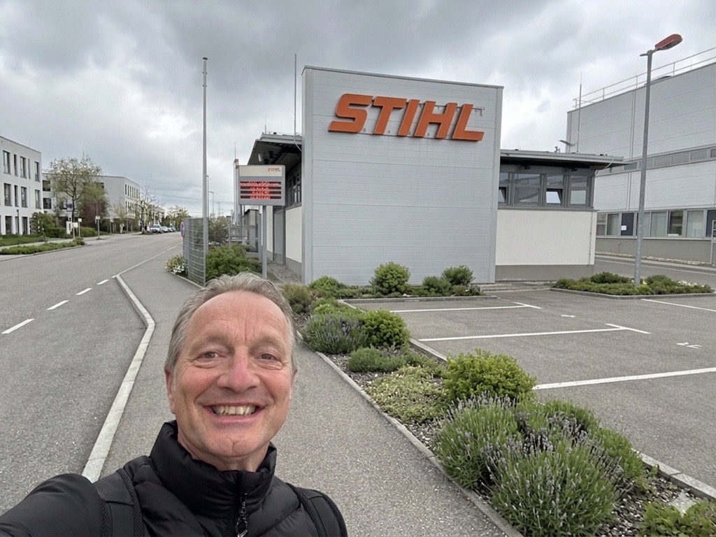 Eine Person mit neutraler Miene steht vor einem grauen STIHL Gebäude bei bewölktem Himmel