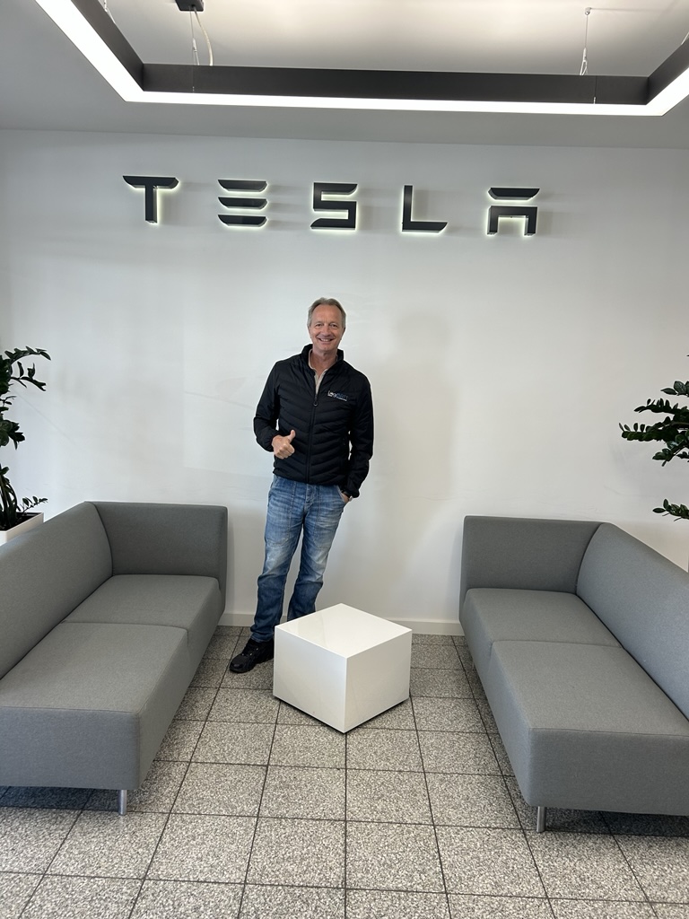 Ein Mann mit Jeans und schwarzer Jacke lächelt vor dem Tesla Logo und zeigt Daumen hoch