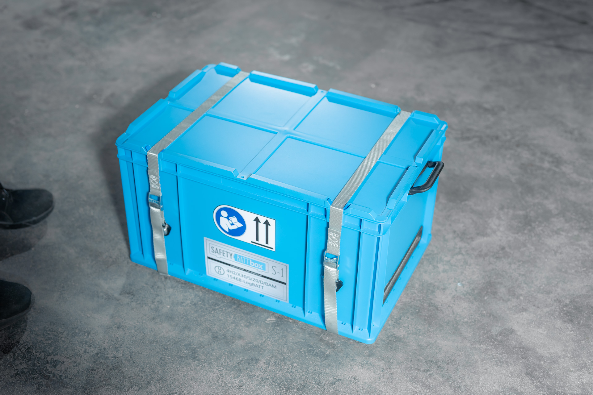 Hellblaue SafetyBATTbox S-1 mit Gurten, neutral, Warnsymbol und Griffen auf Betonboden